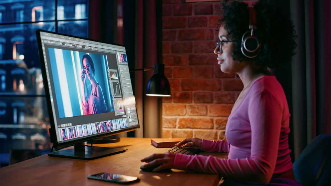 Woman with headphones is editing photos in a photo editing software on a desktop monitor at night.
